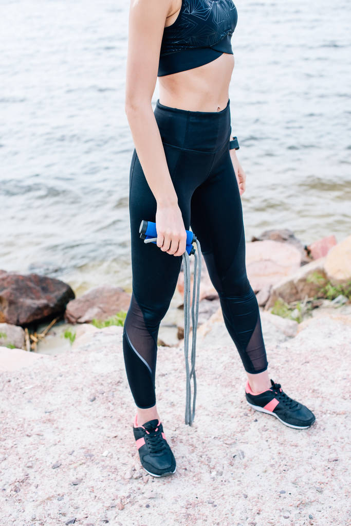 vista cortada de jovem mulher em sportswear segurando pulando corda perto do mar
  - Foto, Imagem
