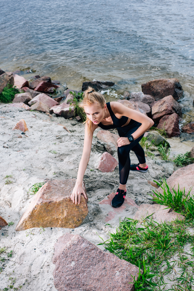 εναέρια θέα της πανέμορφης ξανθιά αθλητική γυναίκα στην αναρρίχηση αθλητικών ειδών κοντά σε πέτρες και θάλασσα - Φωτογραφία, εικόνα