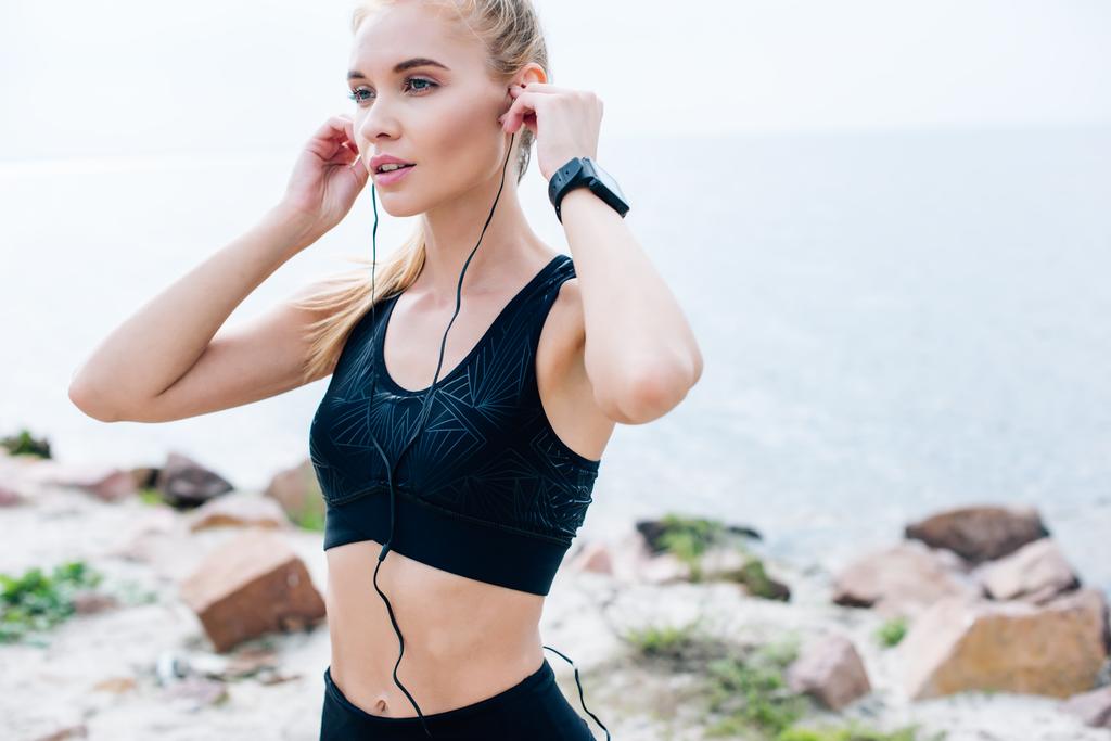 beautiful athletic girl touching earphones and listening music near sea  - Photo, Image