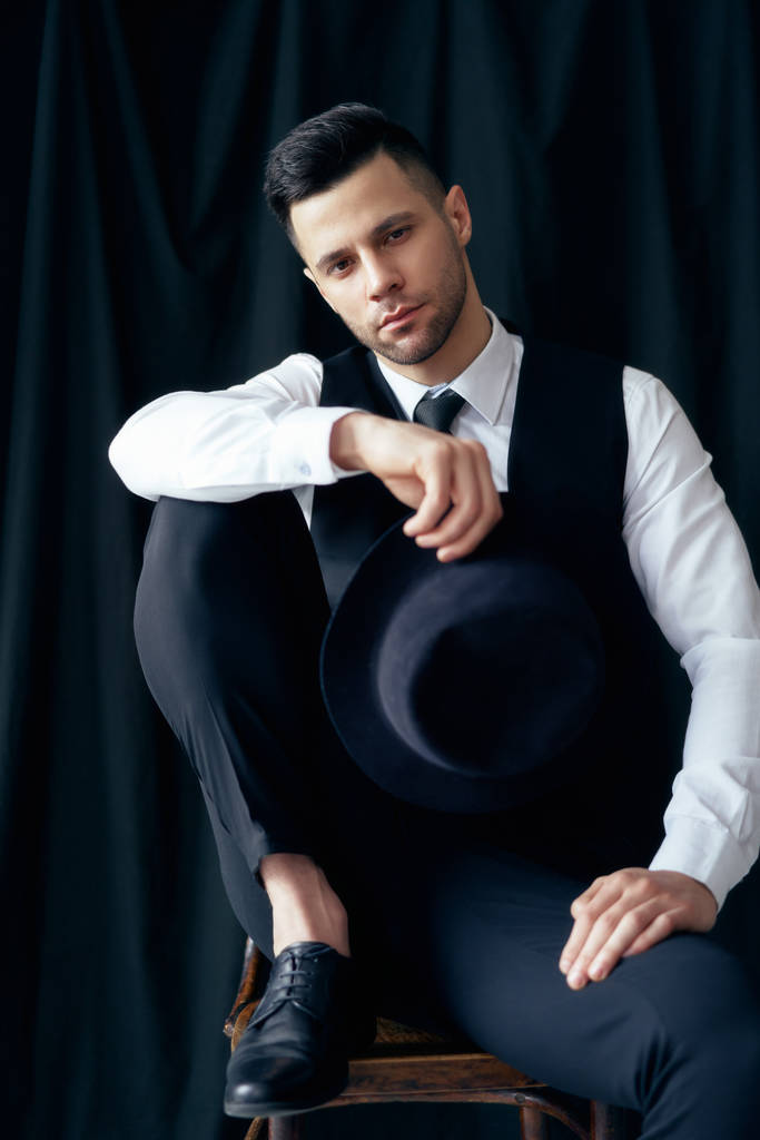 Siyah arka plan üzerinde stüdyopozlama elinde şapka ile zarif takım elbiseli yakışıklı genç adam. Retro tarzı - Fotoğraf, Görsel