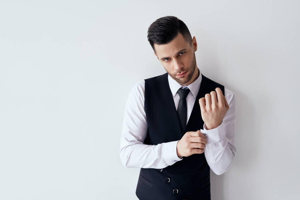 Όμορφος νέος άντρας που ρυθμίζει τα μανίκια του και διορθώνει τις χειροπέδες του ενάντια στο λευκό φόντο με το χώρο αντιγραφής     - Φωτογραφία, εικόνα
