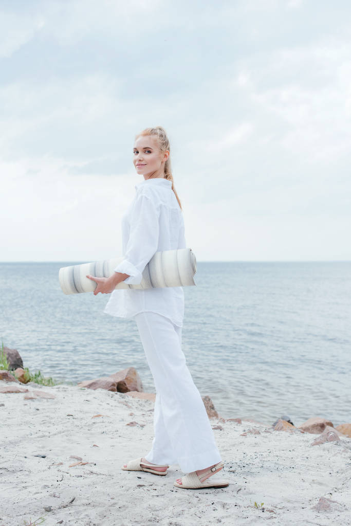sorridente donna bionda in possesso di tappetino yoga e in piedi vicino al mare
  - Foto, immagini