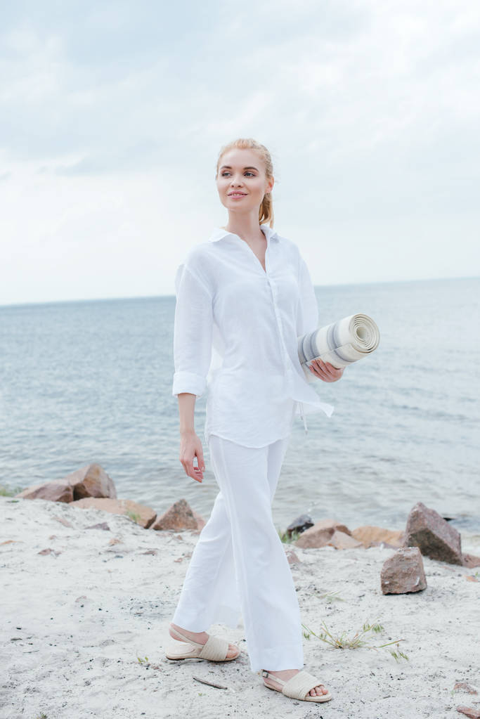 blond kobieta trzyma Mata do jogi i stoi w pobliżu morza  - Zdjęcie, obraz