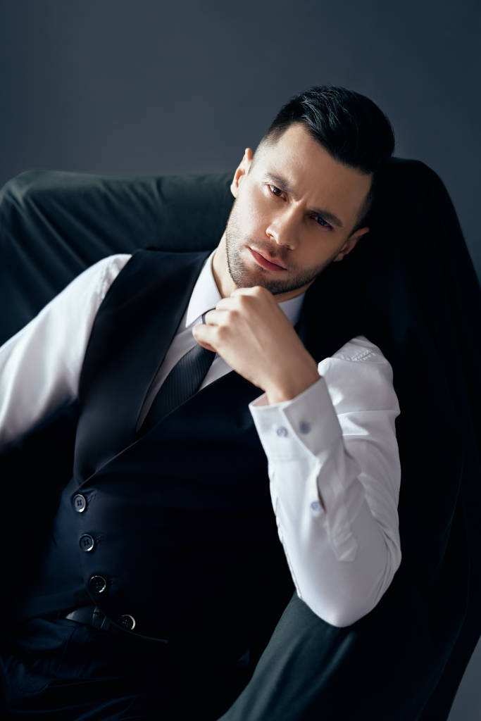 selbstbewusster, nachdenklicher Mann im eleganten Anzug posiert sitzend im Stuhl auf schwarzem Hintergrund - Foto, Bild