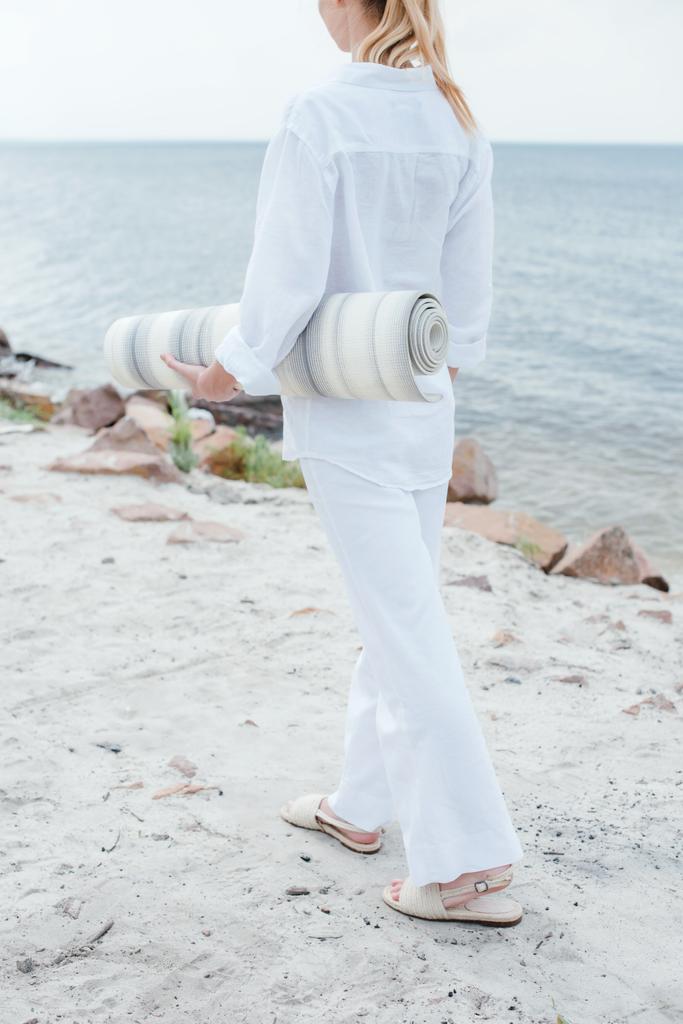 vista cortada da jovem mulher segurando tapete de ioga e de pé perto do mar
  - Foto, Imagem