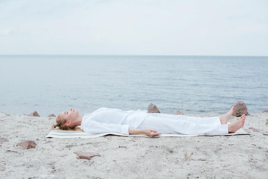 attraente donna bionda con gli occhi chiusi meditando mentre sdraiato sul tappetino yoga vicino al mare
  - Foto, immagini