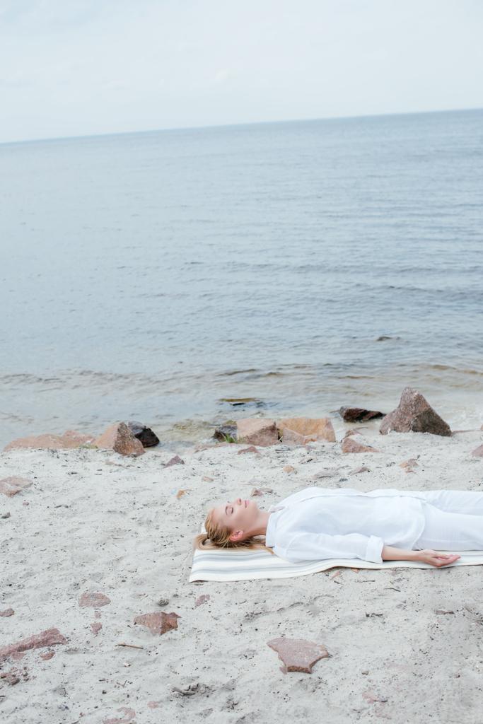 beautiful blonde woman with closed eyes meditating while lying on yoga mat near sea  - Photo, Image