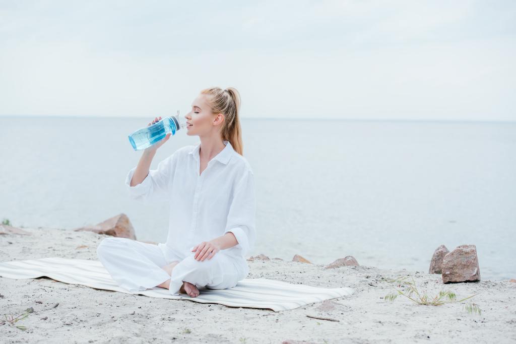 attraktive Frau sitzt auf Yogamatte und hält Sportflasche, während sie Wasser trinkt  - Foto, Bild