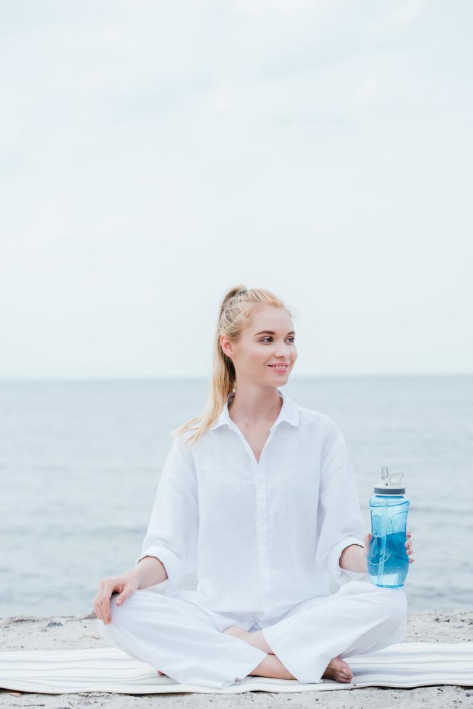 χαρούμενη ξανθιά γυναίκα κρατώντας το μπουκάλι του αθλητισμού και κάθεται στο χαλάκι γιόγκα κοντά στη θάλασσα  - Φωτογραφία, εικόνα