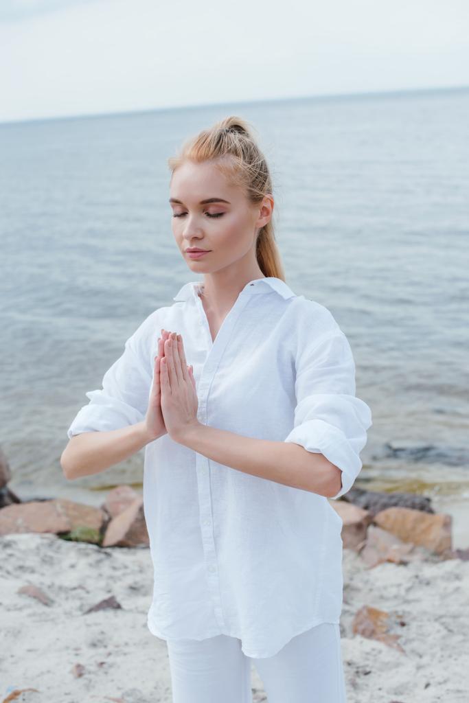 jovem atraente com olhos fechados e mãos de oração perto do mar
  - Foto, Imagem