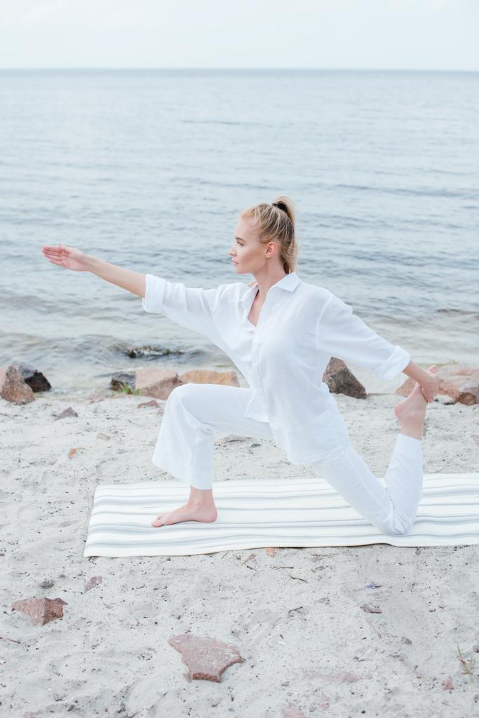 vista lateral de atractiva mujer rubia practicando yoga en estera de yoga cerca del mar
  - Foto, Imagen