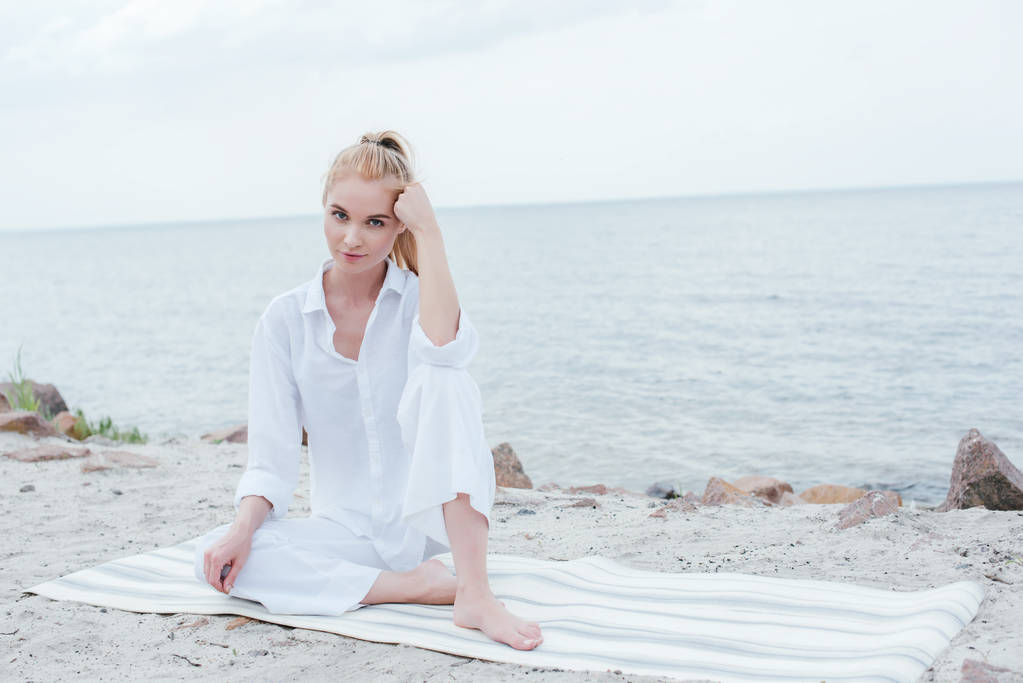 piękna młoda kobieta siedzi na matę do jogi i patrząc na kamerę w pobliżu morza  - Zdjęcie, obraz