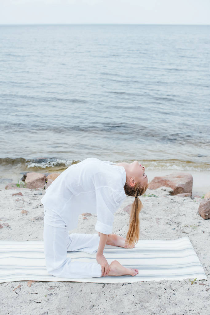 блондинка с закрытыми глазами практикует йогу на коврике для йоги возле моря
  - Фото, изображение