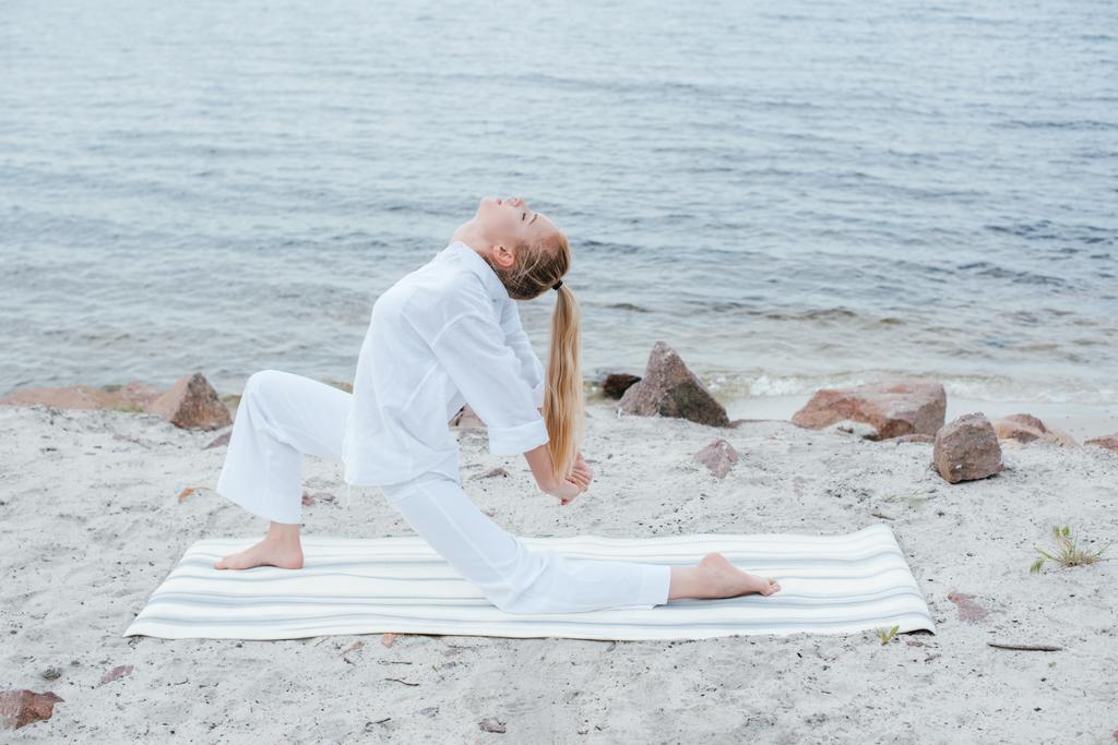 schöne Mädchen mit geschlossenen Augen praktiziert Yoga auf Yogamatte in der Nähe des Meeres  - Foto, Bild