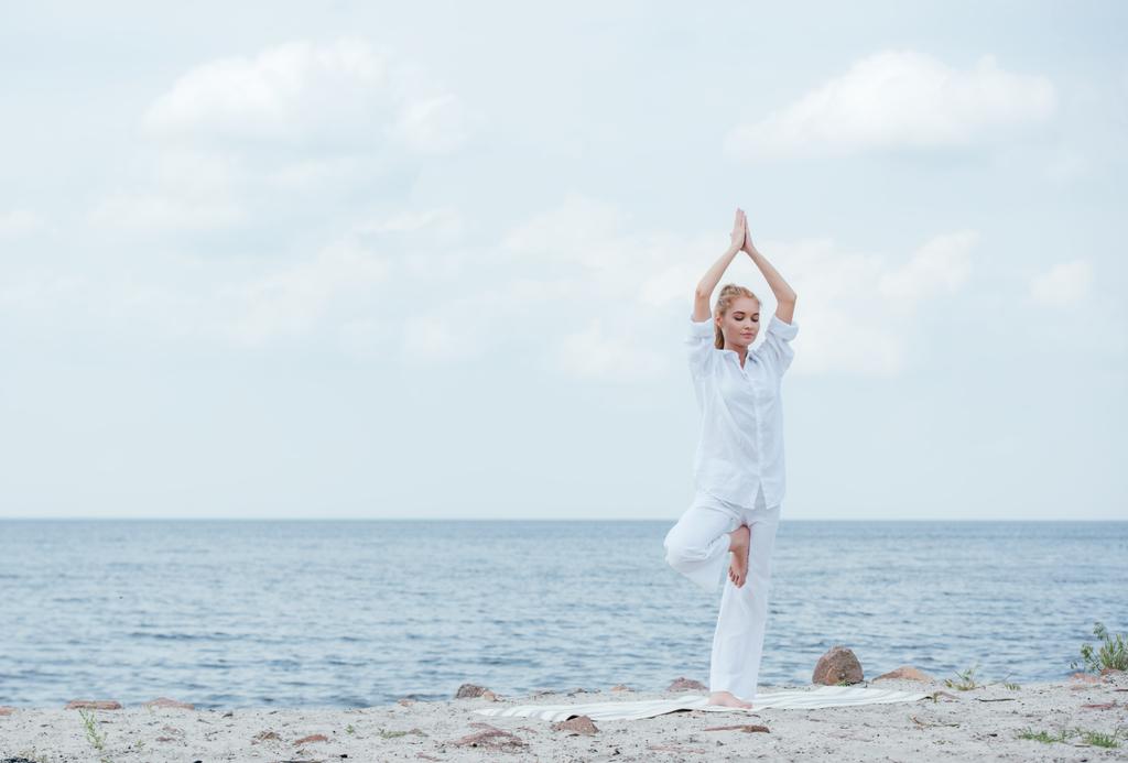 aantrekkelijke blonde vrouw beoefenen van yoga met de handen boven het hoofd in de buurt van zee  - Foto, afbeelding