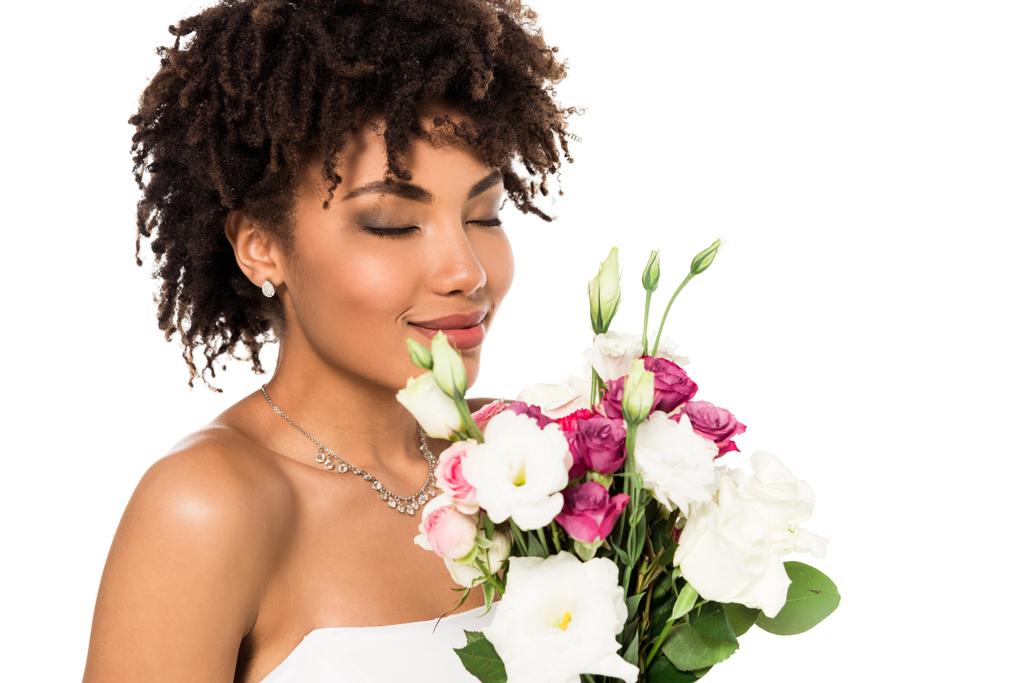 χαρούμενη αφρικανική αμερικανική νύφη κρατώντας μπουκέτο, ενώ μυρίζει λουλούδια απομονώνονται σε λευκό  - Φωτογραφία, εικόνα