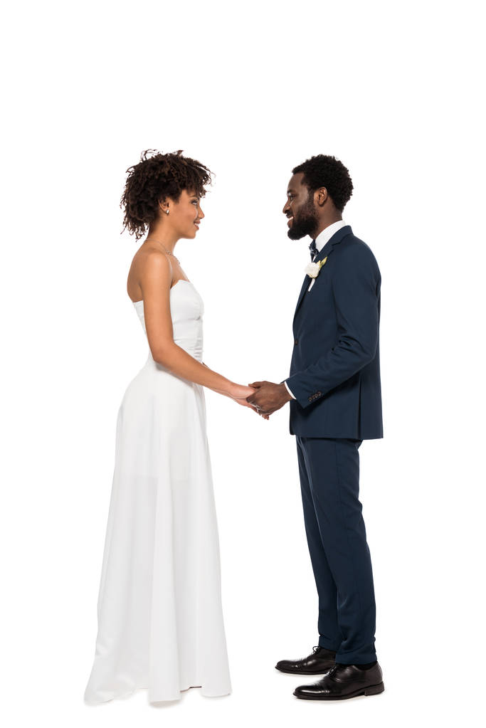 ευτυχισμένος Αφρικανός Αμερικανός γαμπρός και νύφη κρατώντας τα χέρια ενώ κοιτάζοντας ο ένας τον άλλο απομονωμένη σε λευκό  - Φωτογραφία, εικόνα