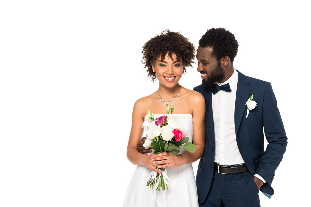 novia afroamericana feliz celebración ramo cerca novio de pie con la mano en el bolsillo aislado en blanco
  - Foto, imagen