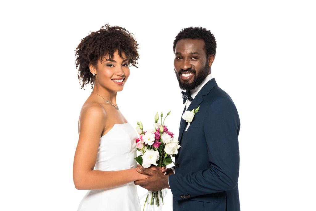 fröhliche afrikanisch-amerikanische Braut mit Blumenstrauß in der Nähe des Bräutigams und Blick in die Kamera isoliert auf weiß  - Foto, Bild