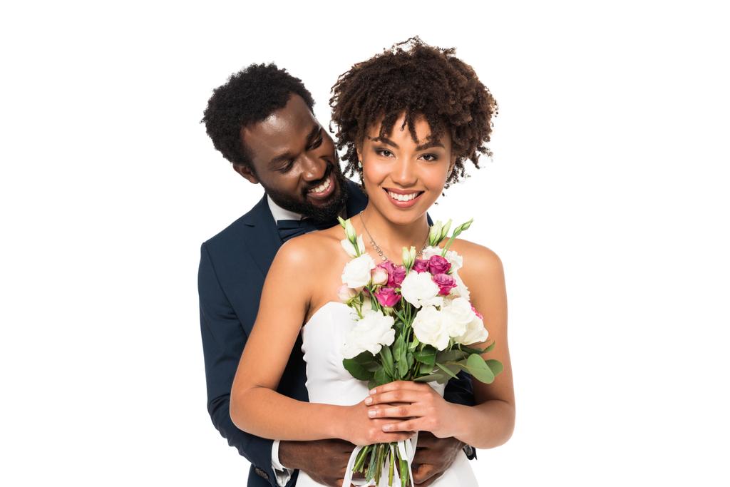 χαμόγελο Αφρικανός Αμερικανός γαμπρός αγκαλιάζοντας ελκυστική νύφη κρατώντας λουλούδια απομονώνονται σε λευκό - Φωτογραφία, εικόνα