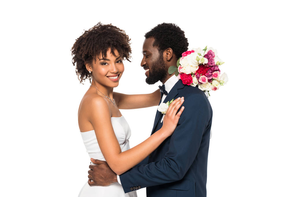 smiling african american bride hugging bridegroom while holding flowers isolated on white - Photo, Image
