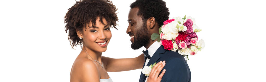 Panoramaaufnahme einer glücklichen afrikanisch-amerikanischen Braut, die den Bräutigam umarmt, während sie Blumen isoliert auf Weiß hält - Foto, Bild
