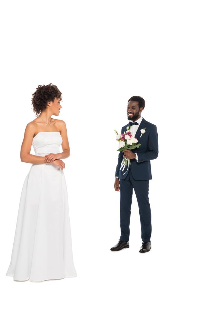 glückliche afrikanisch-amerikanische Braut steht und schaut den bärtigen Bräutigam an, der Blumen isoliert auf weißem Grund hält - Foto, Bild