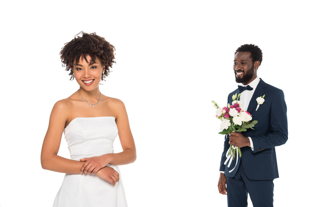 白に隔離された花を持つひげを生やした花婿の近くに立っている幸せなアフリカ系アメリカ人の花嫁の選択的な焦点 - 写真・画像