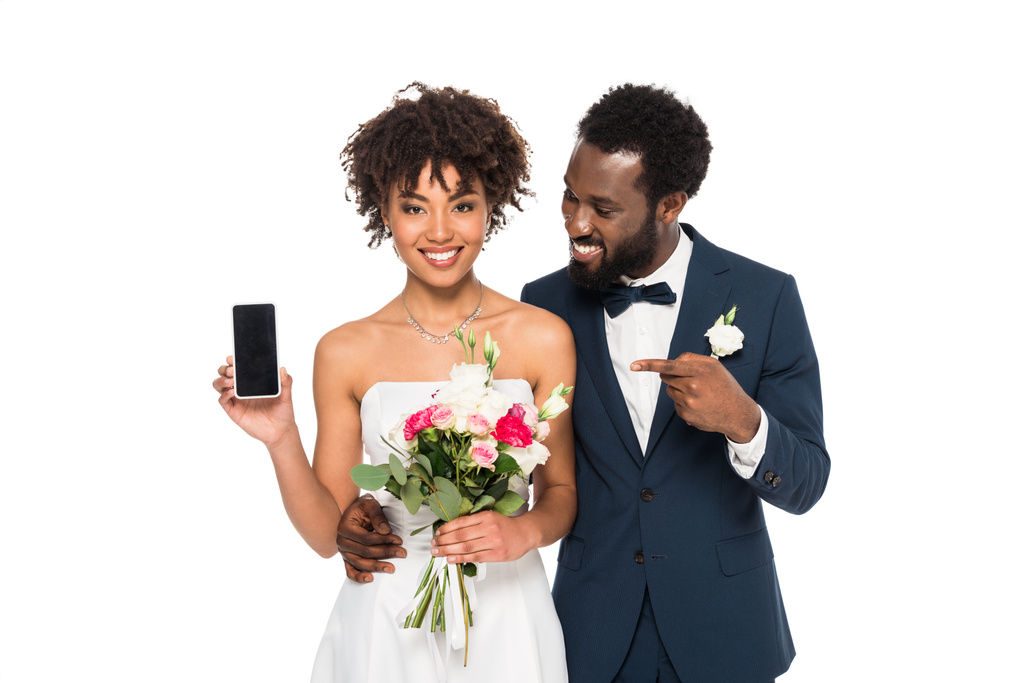 boldog afro-amerikai vőlegény mutató ujját a smartphone üres képernyő közelében menyasszony elszigetelt fehér  - Fotó, kép