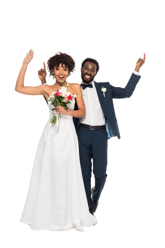 alegre noiva afro-americana de pé com noivo e gestos enquanto segurando flores isoladas em branco
 - Foto, Imagem