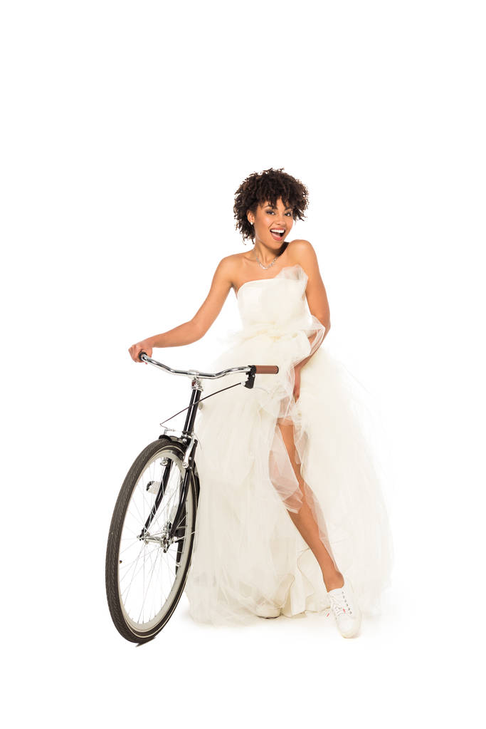 vrolijke Afro-Amerikaanse bruid in trouwjurk rijden fiets geïsoleerd op wit  - Foto, afbeelding