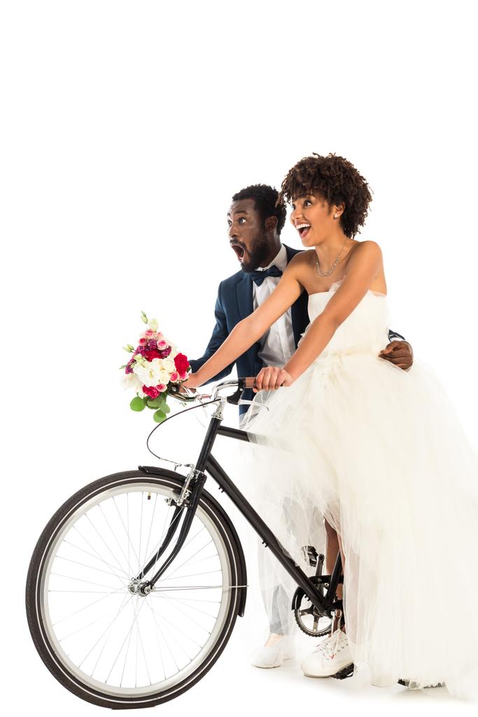 έκπληκτος Αφρικανός Αμερικανός γαμπρός κοντά χαρούμενη νύφη με λουλούδια ιππασία ποδήλατο απομονώνεται σε λευκό  - Φωτογραφία, εικόνα