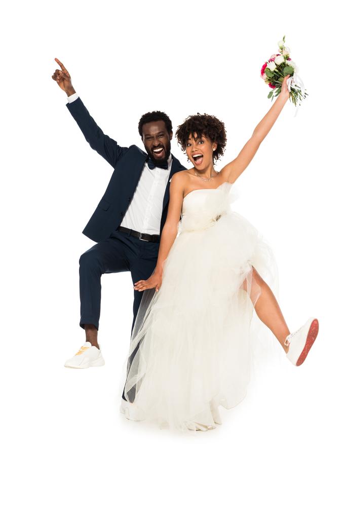 χαρούμενος Αφρικανός Αμερικανός γαμπρός δείχνοντας με το δάχτυλο κοντά ευτυχισμένο νύφη κρατώντας λουλούδια απομονώνονται σε λευκό  - Φωτογραφία, εικόνα