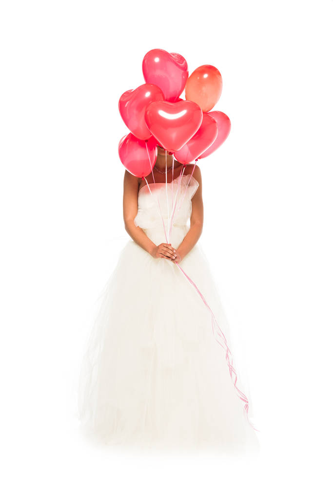 白で隔離されたウェディングドレスに立っている間、ハート型の風船で顔を覆うアフリカ系アメリカ人の花嫁  - 写真・画像