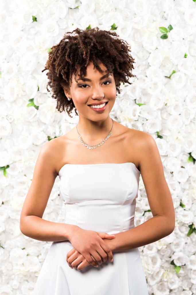 fröhliche afrikanisch-amerikanische Braut blickt in die Kamera, während sie neben Blumen steht  - Foto, Bild