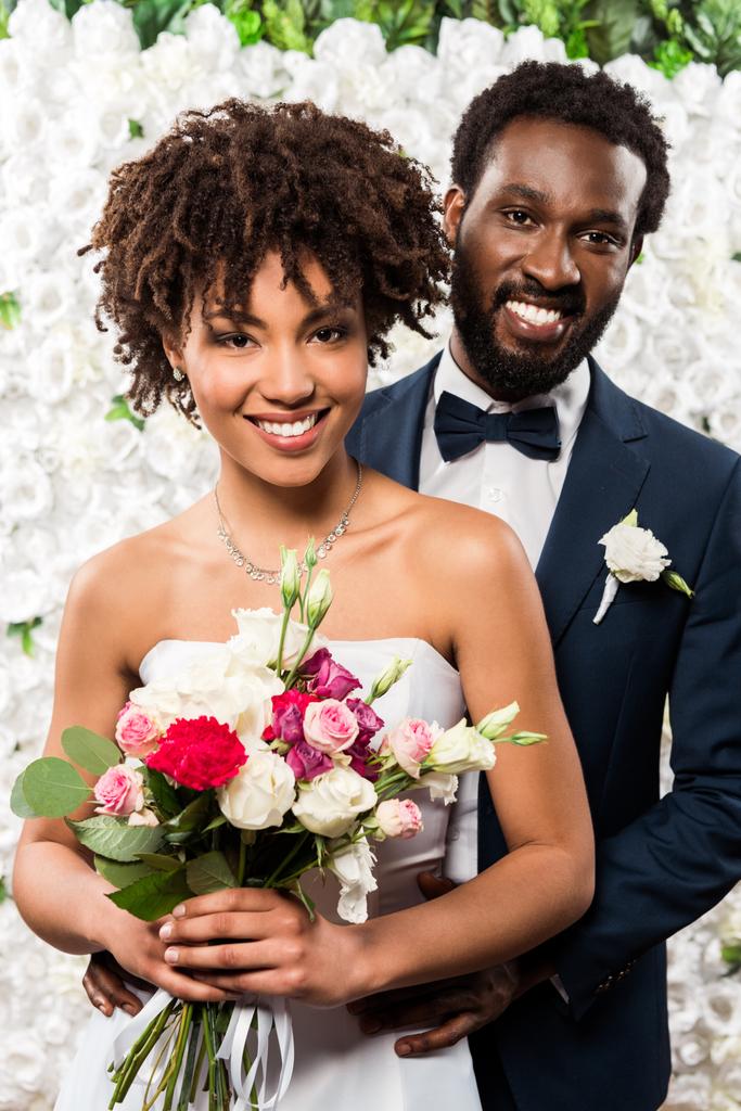 花婿の近くに花と花束を持つ陽気なアフリカ系アメリカ人の花嫁 - 写真・画像