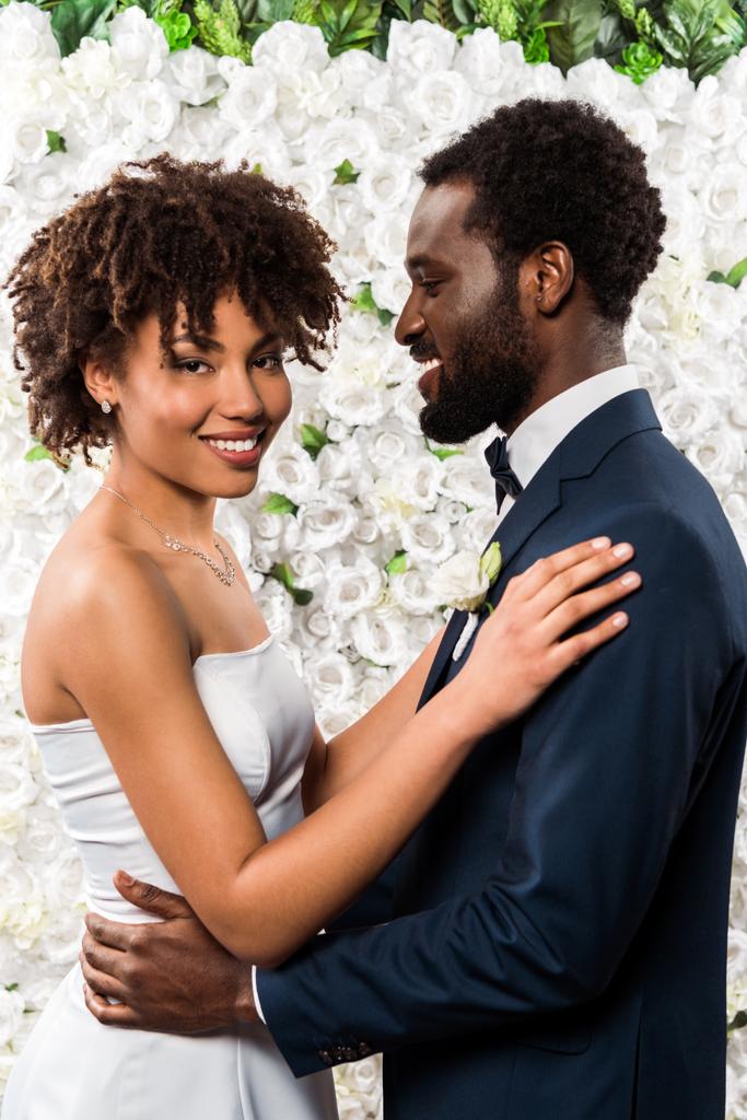 cheerful african american bride hugging happy bridegroom near flowers - Photo, Image