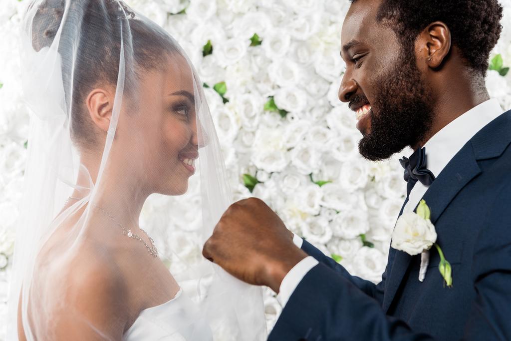 ευτυχισμένος Αφρικανός Αμερικανός γαμπρός αγγίζοντας το λευκό πέπλο και χαμογελά κοντά νύφη και λουλούδια  - Φωτογραφία, εικόνα