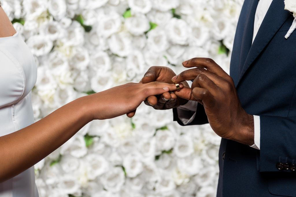 花の近くに花嫁の指に結婚指輪を置くアフリカ系アメリカ人男性のトリミングビュー  - 写真・画像