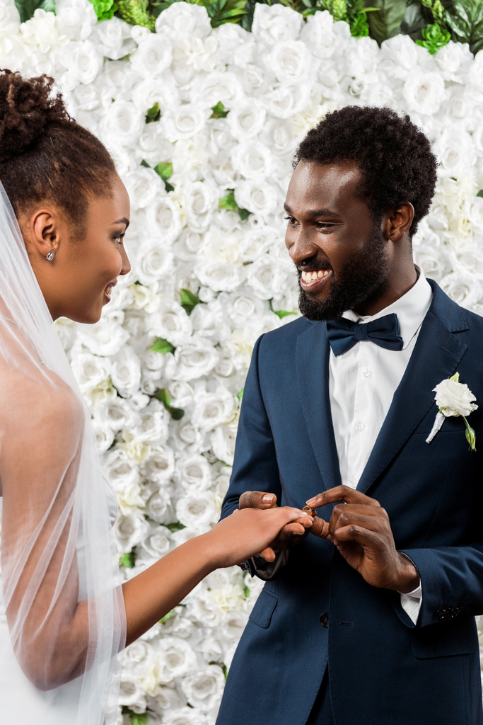 幸せなアフリカ系アメリカ人男性が花の近くに花嫁の指に結婚指輪を置く  - 写真・画像