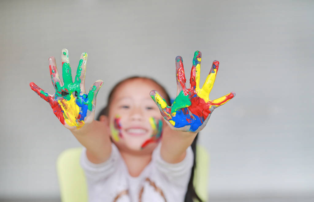 lächelndes kleines fröhliches Mädchen, das seine bunten Hände und Wangen im Kinderzimmer bemalt zeigt. Fokus auf Babyhände. - Foto, Bild