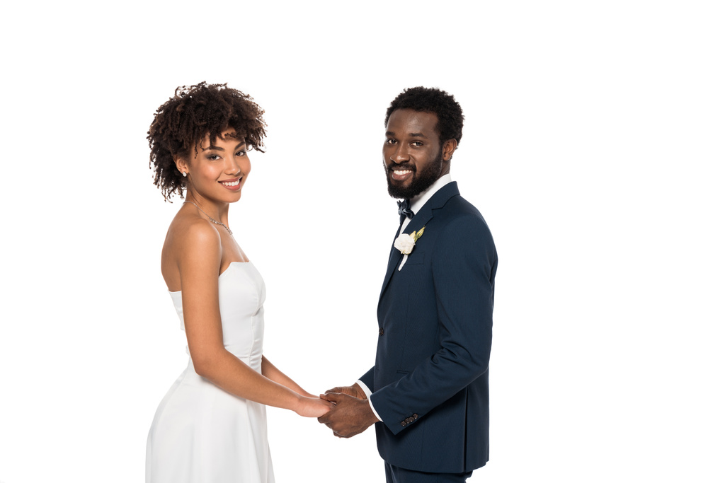 glückliche afrikanisch-amerikanische Braut im Brautkleid und Bräutigam schauen in die Kamera, während sie die Hände isoliert auf weiß halten  - Foto, Bild
