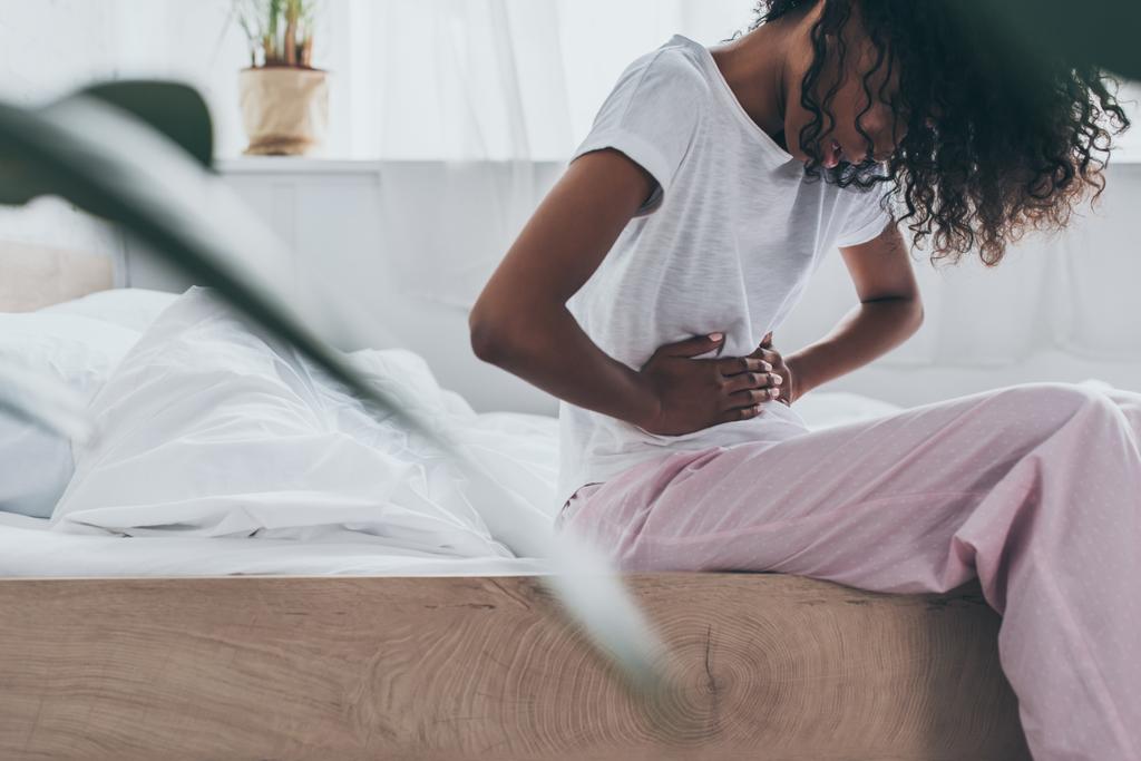 fiatal afro-amerikai nő szenved hasi fájdalom, miközben ül az ágyban - Fotó, kép