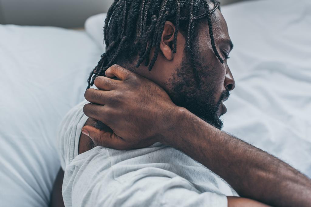 νεαρός Αφροαμερικάνος με κοτσιδάκια που υποφέρει από πόνο στο λαιμό στην κρεβατοκάμαρα. - Φωτογραφία, εικόνα