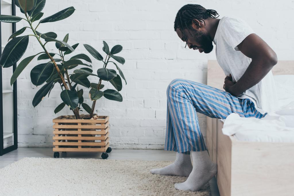 Genç Afrikalı Amerikalı adam yatakta otururken mide ağrısı çekiyor. - Fotoğraf, Görsel