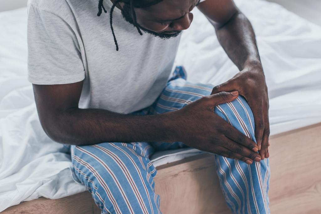 περικομμένη όψη του νεαρού Αφρικανός άνθρωπος που υποφέρει από πόνο στο γόνατο στο υπνοδωμάτιο - Φωτογραφία, εικόνα