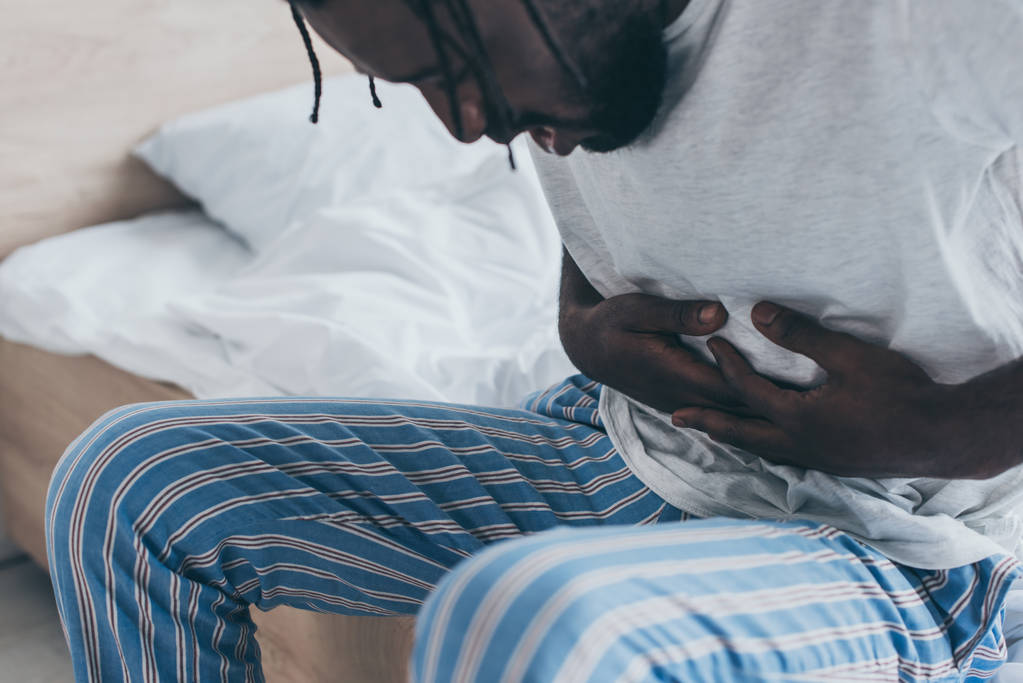 vista parcial del hombre afroamericano que sufre de dolor de estómago en el dormitorio
 - Foto, Imagen