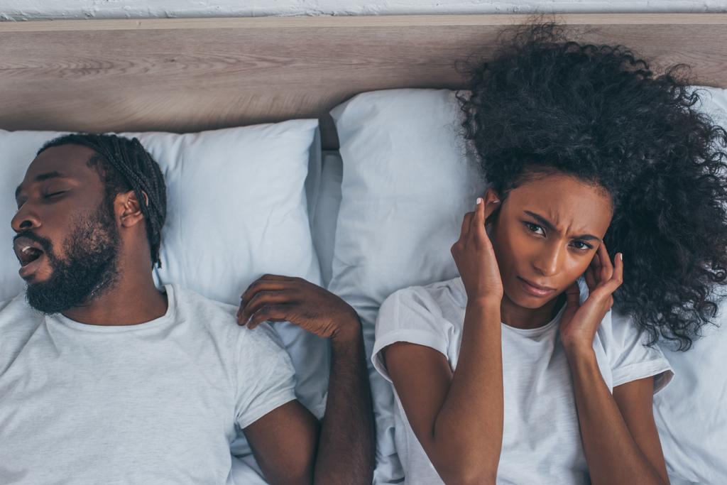 von oben: Afroamerikanerin leidet unter Kopfschmerzen, während sie in der Nähe ihres schnarchenden Ehemannes im Bett liegt - Foto, Bild
