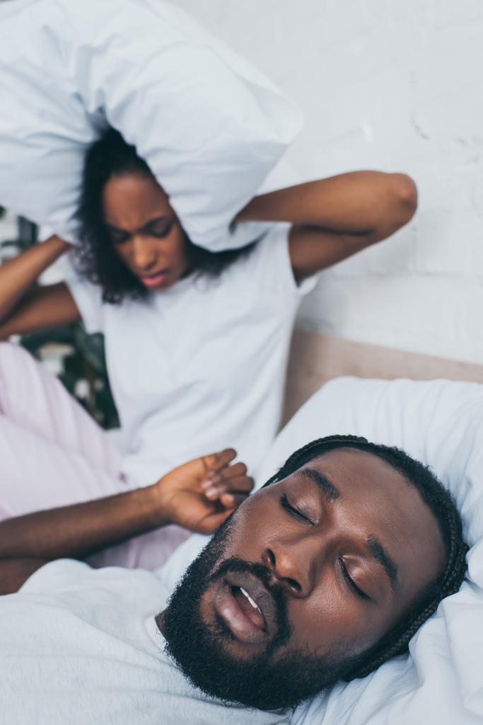いびきをかく夫の近くのベッドに座っている間、枕で頭を覆うアフリカ系アメリカ人女性の選択的な焦点 - 写真・画像