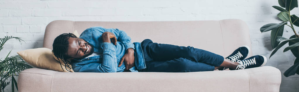 Panoramaaufnahme eines afrikanisch-amerikanischen Mannes, der auf dem Sofa liegend unter Bauchschmerzen leidet - Foto, Bild